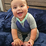 Thiago is registered to the contest to win money with this photo: baby, child, smiling, happy, sitting, blue_blanket, clothing, hands, feet, face, infant, cute, indoors, portrait, wooden_background, cozy, skin, expression, joy, toddler