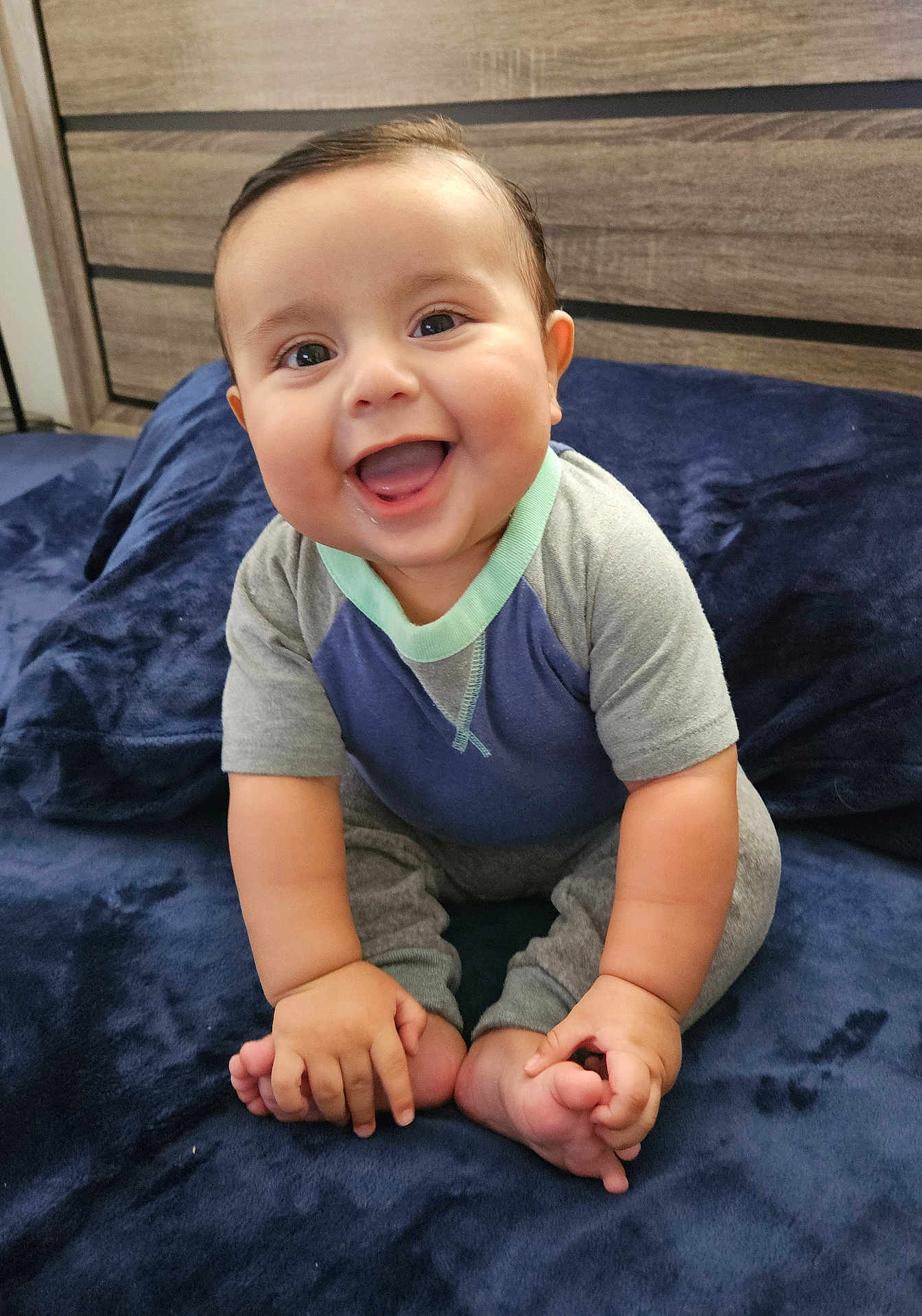 Thiago is registered to the contest to win money with this photo: baby, child, smiling, happy, sitting, blue_blanket, clothing, hands, feet, face, infant, cute, indoors, portrait, wooden_background, cozy, skin, expression, joy, toddler