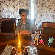 Leilani is registered to the contest to win money with this photo: child, birthday_cake, candle, table, doll, toy_horse, curtains, girl, pigtails, denim_jacket, room, celebration, foil, chocolate_cake, number_candle, wooden_chair, toy_brush, indoor, happy, party