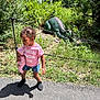 child, doll, pink_shirt, denim_shorts, outdoor, greenery, dinosaur_statue, chain_fence, sunlight, shadow, grass, path, nature, toy, person, summer_clothing, hair_buns, playful, park, daytime