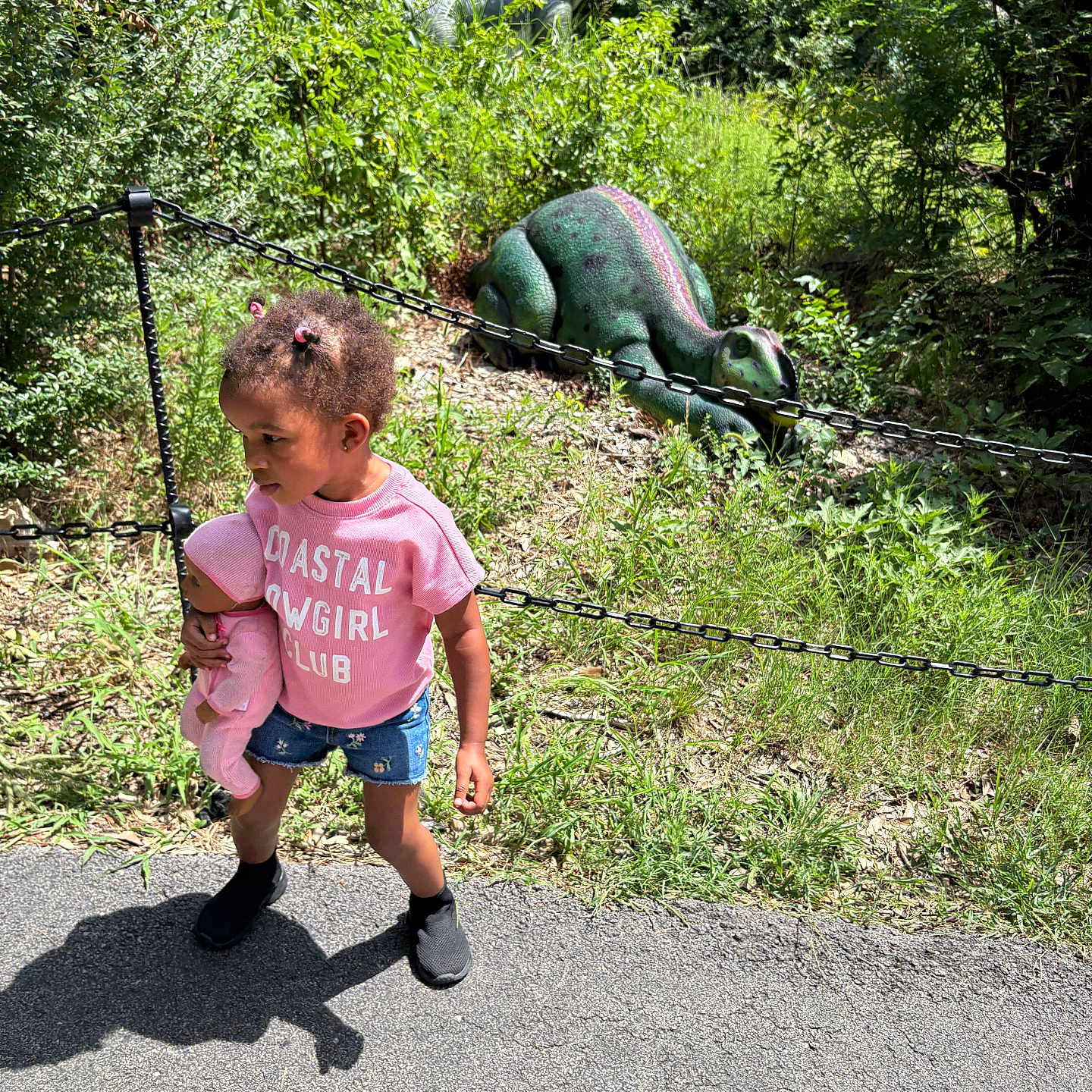 Joyous joined the competition — help win amazing prizes! chain_fence, child, daytime, denim_shorts, dinosaur_statue, doll, grass, greenery, hair_buns, nature, outdoor, park, path, person, pink_shirt, playful, shadow, summer_clothing, sunlight, toy