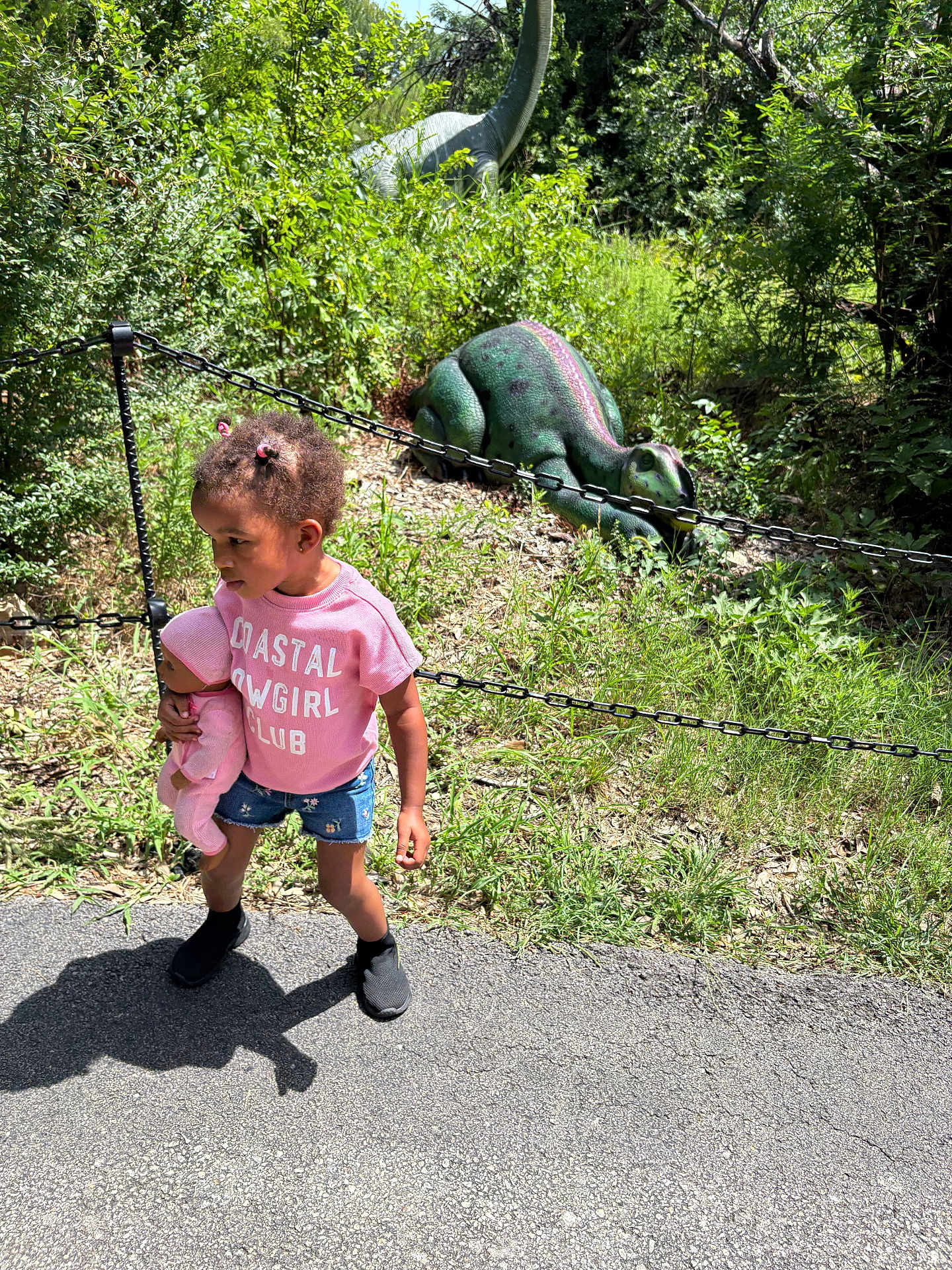 Joyous joined the competition — help win amazing prizes! child, doll, pink_shirt, denim_shorts, outdoor, greenery, dinosaur_statue, chain_fence, sunlight, shadow, grass, path, nature, toy, person, summer_clothing, hair_buns, playful, park, daytime