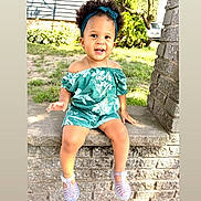Aubree is registered to the contest to win money with this photo: toddler, child, smiling, curly_hair, headband, greenery, outdoor, sunlight, stone_ledge, romper, off_shoulder, shoes, jelly_shoes, necklace, earrings, grass, tree, brick_wall, happy, portrait