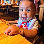 toddler, child, curly_hair, pink_bow, bracelets, bib, restaurant, wooden_table, menu, indoor, portrait, cute, happy, seated, person, closeup, bright_lighting, expression, curious, casual_clothing