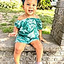 toddler, child, outdoor, smiling, curly_hair, headband, green_clothing, floral_pattern, necklace, stone_ledge, brick_wall, grass, sunlight, happy, shoes, sitting, person, portrait, casual, daylight