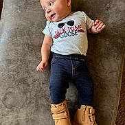 Maverick is registered to the contest to win money with this photo: baby, child, infant, tshirt, boots, pants, smile, face, hand, footwear, clothing, lying_down, curious, indoor, soft_surface, light_skin, short_hair, casual_wear, portrait, cute
