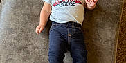 Maverick is registered to the contest to win money with this photo: baby, child, smiling, shirt, text_on_clothing, pants, boots, footwear, lying_down, indoor, carpet, fabric, cute, infant, person, portrait, happy, casual_clothing, light_hair, small_child
