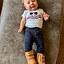 baby, child, smiling, shirt, text_on_clothing, pants, boots, footwear, lying_down, indoor, carpet, fabric, cute, infant, person, portrait, happy, casual_clothing, light_hair, small_child