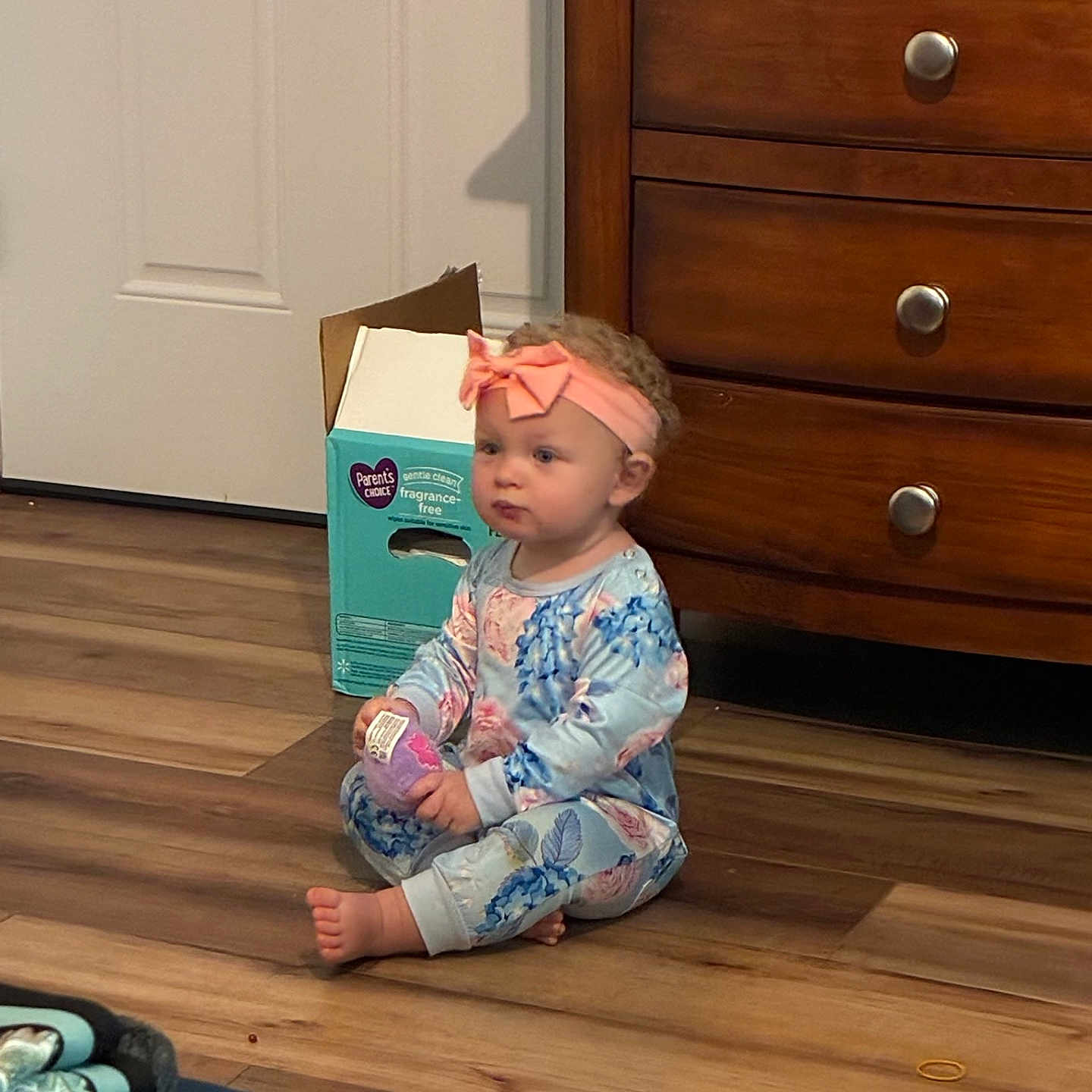Aurora is registered to the contest to win money with this photo: baby, child, toddler, sitting, portrait, pink_bow, headband, floral_clothing, dress, hardwood_floor, dresser, furniture, tissue_box, indoors, home, door, books, barefoot, curly_hair, cute