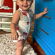 Walker is registered to the contest to win money with this photo: toddler, child, smiling, standing, indoor, rug, dresser, blue_furniture, red_cushion, white_chair, floor, home_interior, clothing, sleeveless, baby, hand_on_furniture, happy, cute, portrait, person