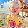 child, smiling, outdoor, sunlight, dress, blonde_hair, cartoon_character, building, deck, blue_sky, happy, person, grass, concrete, daylight, cute, toddler, clothing, portrait, young