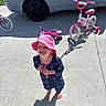Amirah is registered to the contest to win money with this photo: child, toddler, pink_hat, blue_dress, barefoot, bicycle, sidewalk, car, outdoor, sunlight, shadow, smiling, happy, playtime, concrete, daytime, vehicle, helmet, toy, recreation