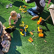 Ryder joined the competition — help win amazing prizes! toddler, adult, toy_tools, grass, outdoor, playtime, yellow, black_shirt, jeans, shoes, toy_trucks, sunlight, garden, plastic_tools, hat, decorative_statue, cinder_blocks, child_playing, person, casual_clothing