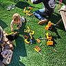 toddler, adult, toy_tools, grass, outdoor, playtime, yellow, black_shirt, jeans, shoes, toy_trucks, sunlight, garden, plastic_tools, hat, decorative_statue, cinder_blocks, child_playing, person, casual_clothing