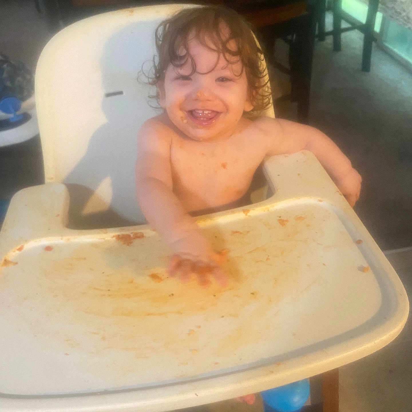 Chase is registered to the contest to win money with this photo: toddler, high_chair, messy, food, smiling, child, indoor, happy, face, curly_hair, tray, furniture, table, seat, home, baby, eating, dining_room, person, light