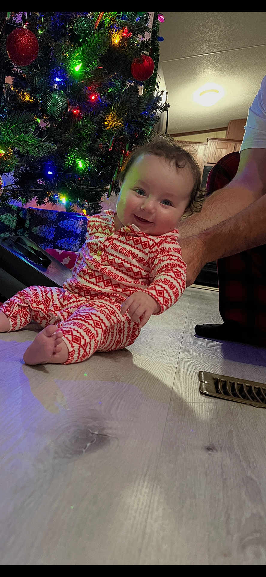 Sariah joined the competition — help win amazing prizes! baby, child, christmas_tree, holiday, pajamas, indoor, floor, smiling, person, adult_arm, decorations, ornaments, lights, festive, wood_floor, home, cozy, seasonal, celebration, happy
