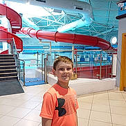 Alfie joined the competition — help win amazing prizes! boy, ceiling, child, door, facility, glass_panel, indoor, lighting, orange_tshirt, person, pool_area, railing, recreation, safety_sign, short_hair, smiling, stairs, swimming_pool, tile_floor, water_slide