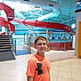 boy, ceiling, child, door, facility, glass_panel, indoor, lighting, orange_tshirt, person, pool_area, railing, recreation, safety_sign, short_hair, smiling, stairs, swimming_pool, tile_floor, water_slide