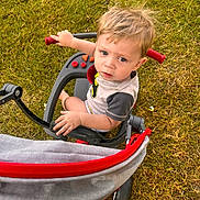 Mason is registered to the contest to win money with this photo: toddler, child, tricycle, outdoor, grass, toy, vehicle, blonde_hair, curious, playing, summer, sunlight, hand, footwear, casual_clothing, expression, seat, handlebar, wheel, person