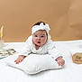 baby, infant, headband, white_clothing, pillow, white_pillow, lying_down, indoor, soft_background, beige_background, magazine, wooden_sign, six_months, cute, chubby_cheeks, child, portrait, cozy, curious, hands