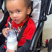 Nolan joined the competition — help win amazing prizes! baby, black_straps, casual_clothing, child, curly_hair, cute, expression, floor_tiles, holding, indoor, looking_up, milk_bottle, person, plastic_bottle, portrait, red_shirt, seated, stroller, toddler, young_child