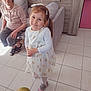 child, girl, elderly_woman, balloon, white_dress, pink_shoes, hair_clip, smile, indoor, tile_floor, furniture, sofa, cabinet, bag, woman, person, happy, home, casual, portrait