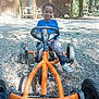 child, smiling, go_kart, outdoor, gravel, trees, wooden_shed, toy_vehicle, happy, casual_clothing, blue_shirt, sneakers, play, daylight, nature, fun, person, young, recreation, park