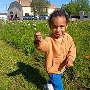 Kaydine participe au concours pour gagner de l'argent avec cette photo : child, toddler, flower, grass, wildflowers, hoodie, blue_pants, sneakers, smile, outdoor, sunny, nature, greenery, curly_hair, parked_cars, building, sidewalk, road, daytime, joyful