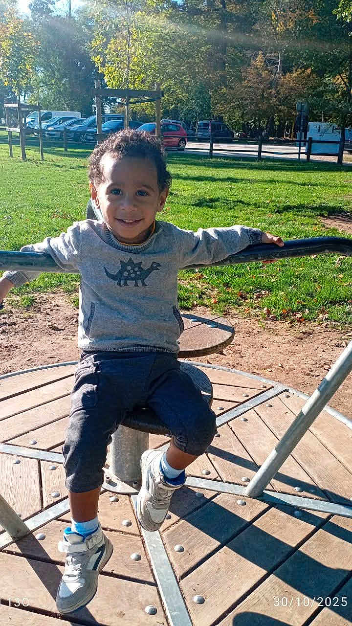 Kaydine participe au concours pour gagner de l'argent avec cette photo : child, smiling, playground, merry_go_round, outdoor, park, grass, trees, sneakers, curly_hair, casual_clothing, sunlight, daytime, happy, person, recreation, fun, young, boy, nature