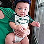 Jasper is registered to the contest to win money with this photo: baby, child, person, cap, blue_cap, green_shirt, window, hand, holding, indoor, natural_light, curious_expression, face, skin, clothing, sitting, closeup, portrait, infant, toddler