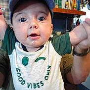 Jasper is registered to the contest to win money with this photo: baby, child, cap, drool, shirt, green, white, person, indoor, bookshelf, furniture, hands, smile, face, curious, joy, holding, casual, cute, young