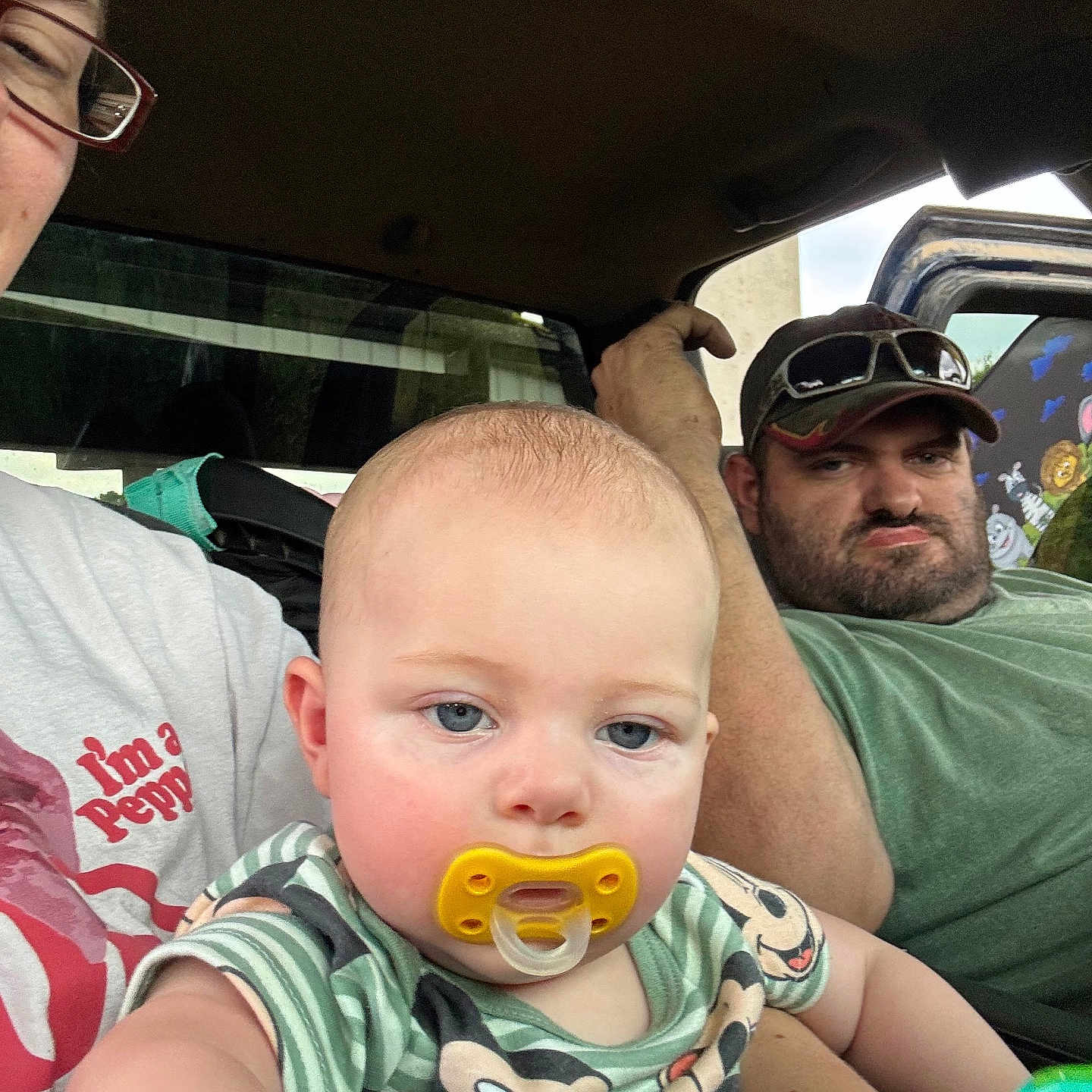 Bobby is registered to the contest to win money with this photo: baby, pacifier, man, woman, glasses, cap, vehicle, window, mural, child, portrait, casual, clothing, indoor, person, seat, green_shirt, striped_shirt, blue_eyes, facial_expression