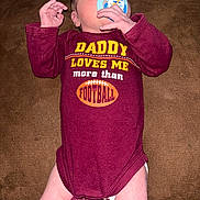 Bobby is registered to the contest to win money with this photo: baby, infant, newborn, pacifier, onesie, maroon_clothing, slogan_on_clothing, text_on_shirt, football_graphic, lying_down, couch, sofa, hand, legs, skin, portrait, closeup, cute, indoor, parental_message