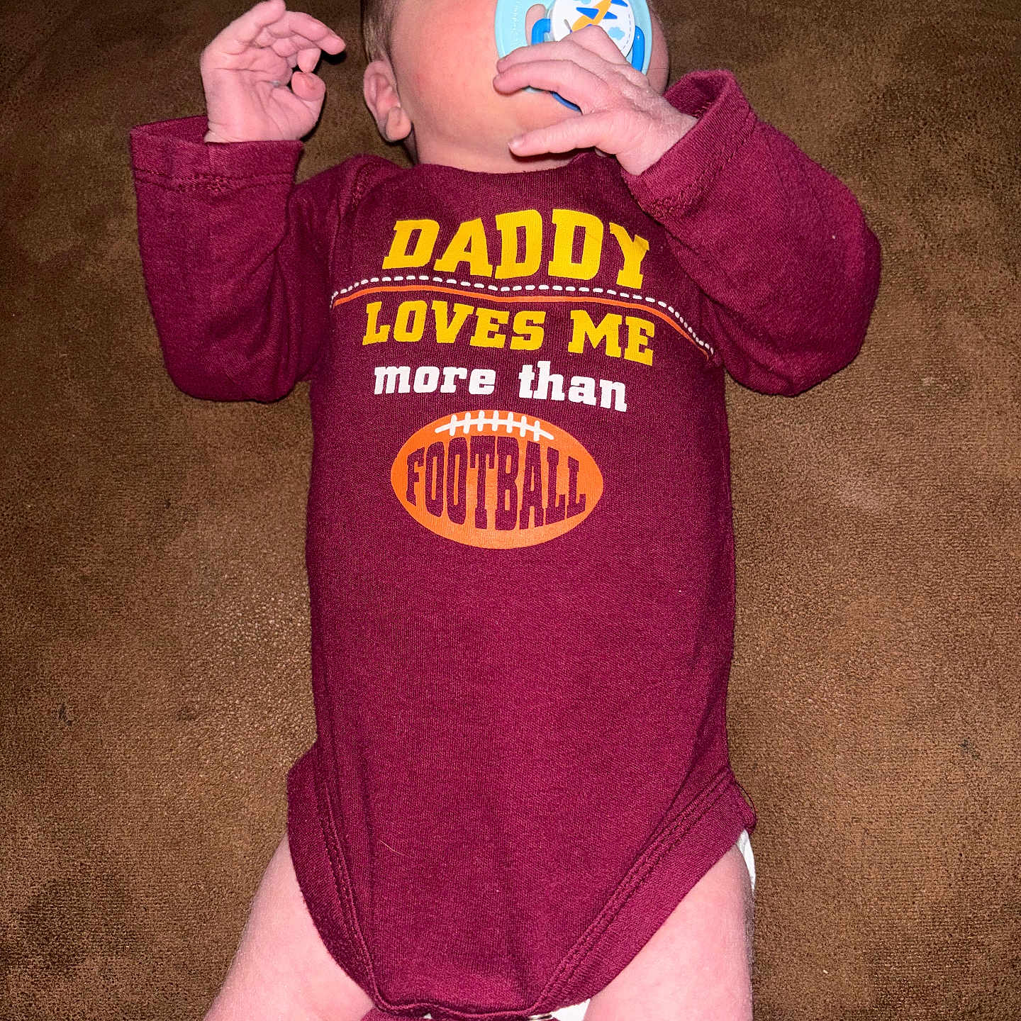 Bobby is registered to the contest to win money with this photo: baby, closeup, couch, cute, football_graphic, hand, indoor, infant, legs, lying_down, maroon_clothing, newborn, onesie, pacifier, parental_message, portrait, skin, slogan_on_clothing, sofa, text_on_shirt
