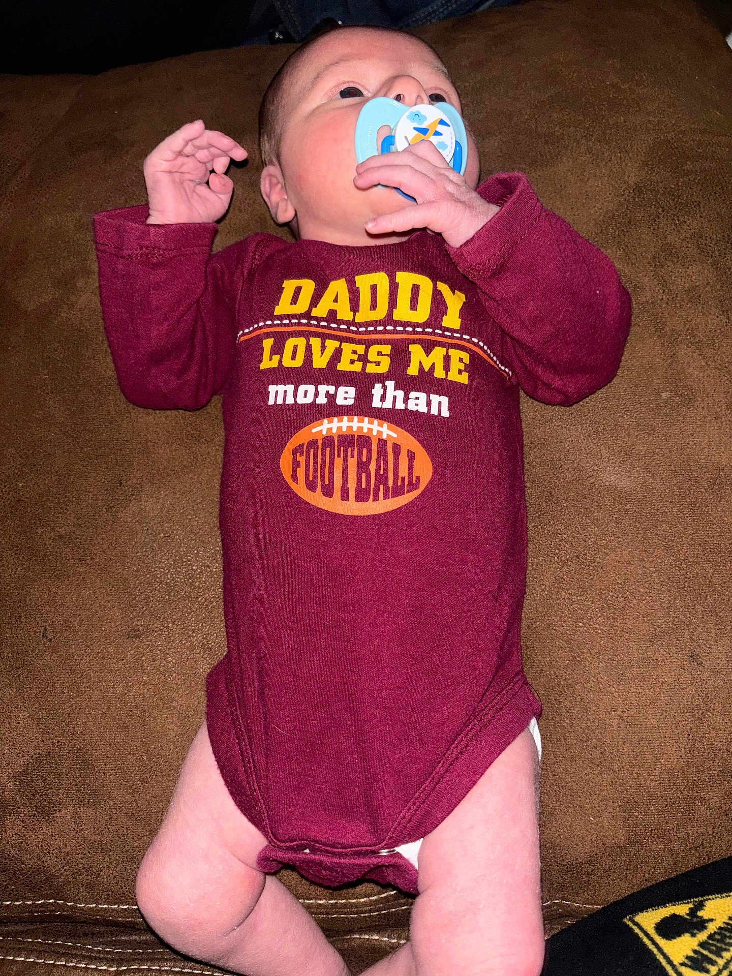 Bobby is registered to the contest to win money with this photo: baby, infant, newborn, pacifier, onesie, maroon_clothing, slogan_on_clothing, text_on_shirt, football_graphic, lying_down, couch, sofa, hand, legs, skin, portrait, closeup, cute, indoor, parental_message