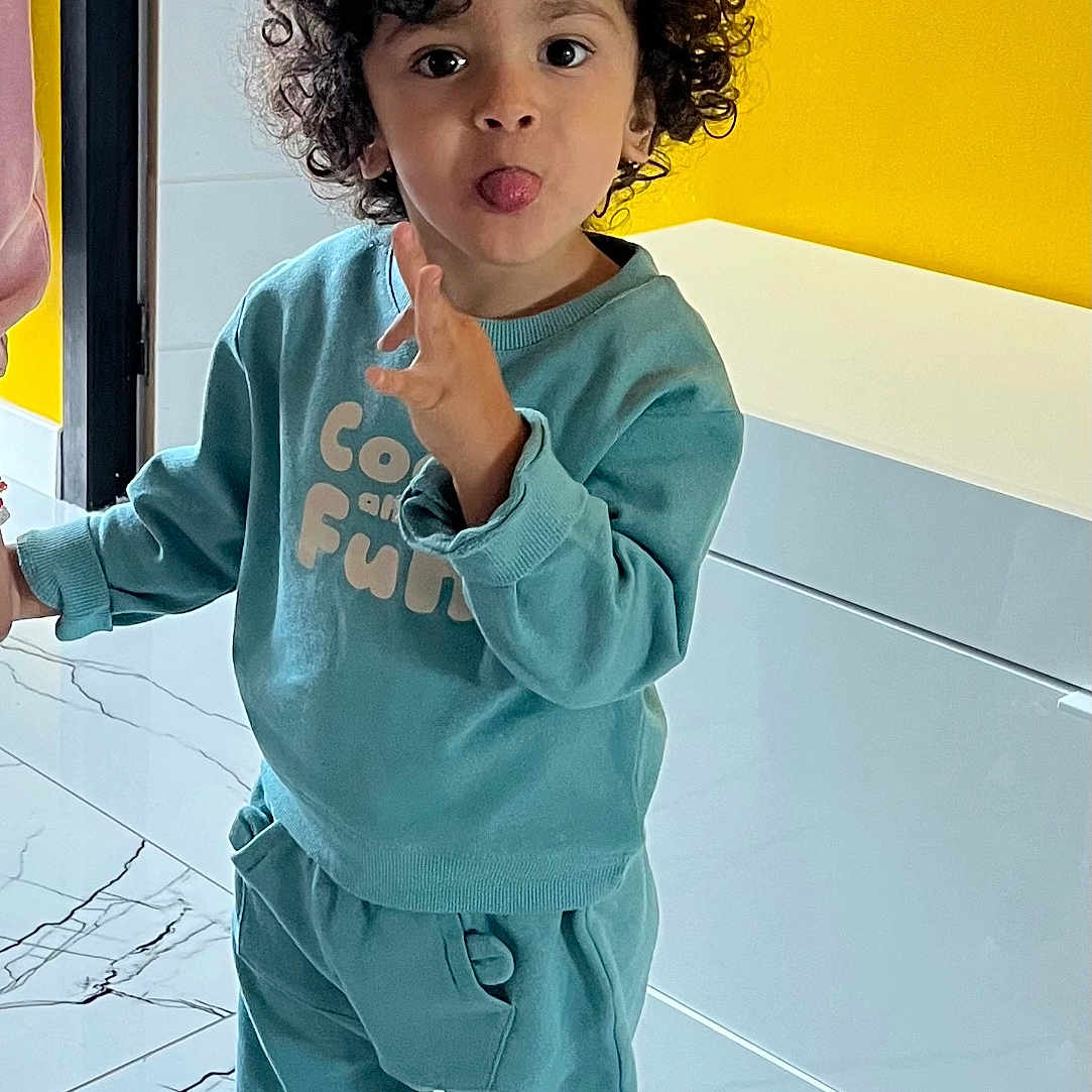 Noha a rejoint le concours — aidez-le/la à gagner de superbes lots ! child, toddler, curly_hair, playful, indoor, yellow_wall, white_cabinet, teal_outfit, sneakers, tongue_out, marvel_shoes, smiling, happy, person, floor, home, casual_clothing, cute, fun, standing