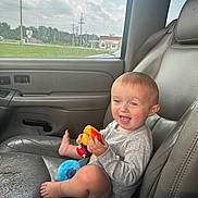 Brooklyn is registered to the contest to win money with this photo: toddler, child, car_interior, car_seat, toy, smiling, happy, baby, window, seatbelt, gray_leather, foot, clothing, daylight, sky, clouds, grass, vehicle, portrait, sitting