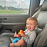 toddler, child, car_interior, car_seat, toy, smiling, happy, baby, window, seatbelt, gray_leather, foot, clothing, daylight, sky, clouds, grass, vehicle, portrait, sitting