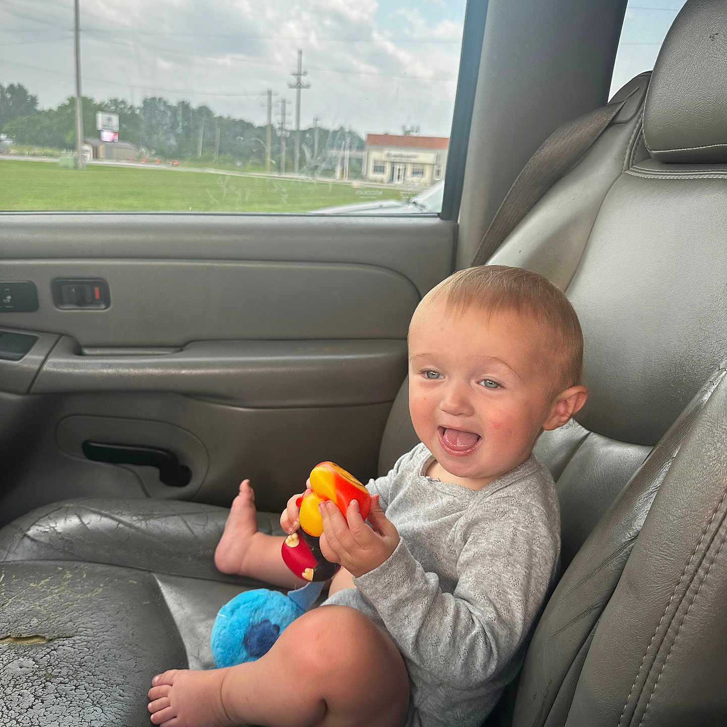 Brooklyn is registered to the contest to win money with this photo: toddler, child, car_interior, car_seat, toy, smiling, happy, baby, window, seatbelt, gray_leather, foot, clothing, daylight, sky, clouds, grass, vehicle, portrait, sitting