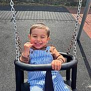 Sebastian joined the competition — help win amazing prizes! child, toddler, swing, playground, smiling, happy, outdoor, daylight, clothing, overalls, brown_hoodie, play, fun, metal_chains, black_swing, stone_wall, asphalt_ground, cute, joyful, person