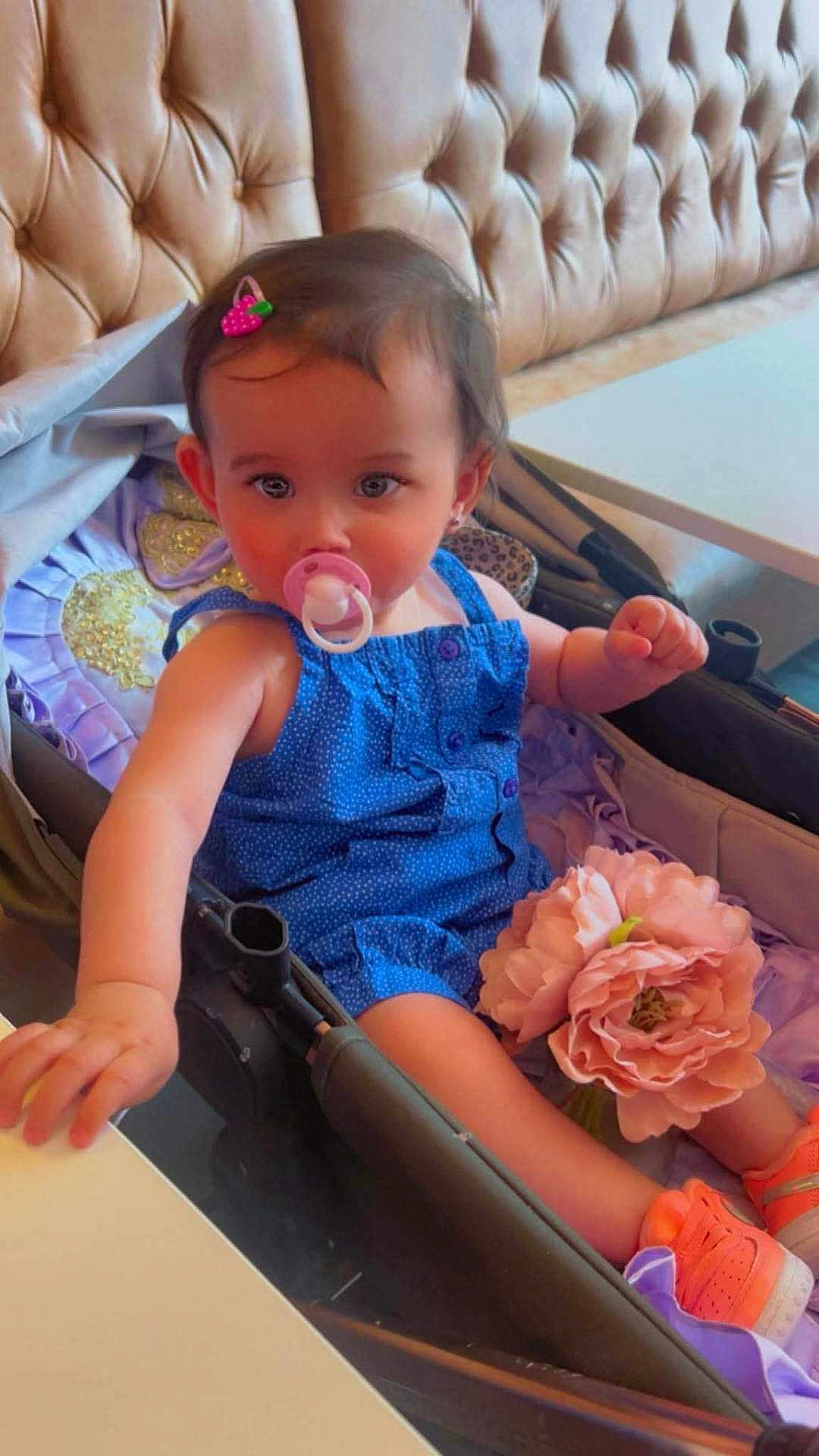 Asiye is registered to the contest to win money with this photo: toddler, child, pacifier, blue_outfit, pink_hair_clip, stroller, flower, orange_shoes, leather_cushion, indoor, cute, baby, portrait, sitting, table, hand, face, head, furniture, soft_lighting