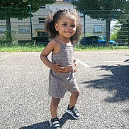 Neliyah a rejoint le concours — aidez-le/la à gagner de superbes lots ! child, outdoor, sunny, smile, curly_hair, checkered_outfit, sneakers, black_shoes, asphalt, fence, trees, building, shadow, grass, person, casual_clothing, daylight, playful, young_child, happy