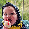 Ezekiel is registered to the contest to win money with this photo: baby, batman, chewing, child, close_up, greenery, hand, happy, hoodie, infant, outdoor, playful, portrait, sitting, smiling, striped_clothing, teething_toy, toy, window, yellow_clothing