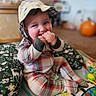 baby, blurred_background, child, colorful, cozy, cushion, finger_biting, happy, hat, indoors, infant, orange_ball, patterned_fabric, plaid_clothing, play_mat, playroom, portrait, sitting, smiling, toys