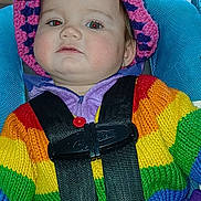Laylani is registered to the contest to win money with this photo: child, toddler, car_seat, rainbow_sweater, knit_hat, seatbelt, indoors, curious, baby, colorful_clothing, face, person, portrait, sitting, wooden_furniture, flooring, red_button, purple_clothing, blue_seat, toy
