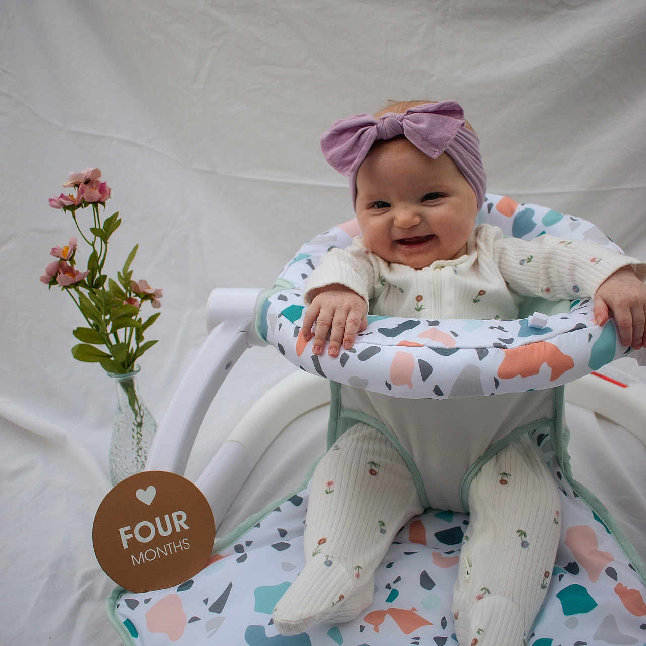Aurora joined the competition — help win amazing prizes! baby, baby_chair, child, cute, floral_print, four_months, happy, headband, indoor, infant, onesie, patterned_fabric, pink_flowers, portrait, purple_bow, seated, smiling, toddler, vase, white_background
