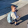 child, boy, backpack, toy, outdoor, sidewalk, sunlight, shadow, happy, casual_clothing, shorts, sneakers, playful, daylight, pavement, concrete_wall, expression, walking, small_toy_car, urban