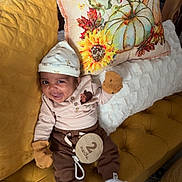 Leon is registered to the contest to win money with this photo: baby, infant, couch, pillow, pumpkin, sunflower, hat, mittens, socks, wooden_sign, 2_months, smiling, tufted_couch, yellow, brown_clothing, striped_shirt, cozy, indoor, carpet, person