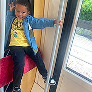 Ibrahima a rejoint le concours — aidez-le/la à gagner de superbes lots ! child, boy, smiling, blue_jacket, yellow_tshirt, black_pants, black_and_white_sneakers, pole, red_seat, window, indoor, public_transport, playful_pose, hand_gesture, happy, person, casual_clothing, daylight, curtain, greenery_outside