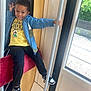 child, boy, smiling, blue_jacket, yellow_tshirt, black_pants, black_and_white_sneakers, pole, red_seat, window, indoor, public_transport, playful_pose, hand_gesture, happy, person, casual_clothing, daylight, curtain, greenery_outside